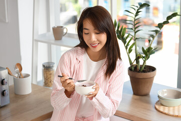 Pretty young Asian woman in pajamas eating tasty yogurt at home