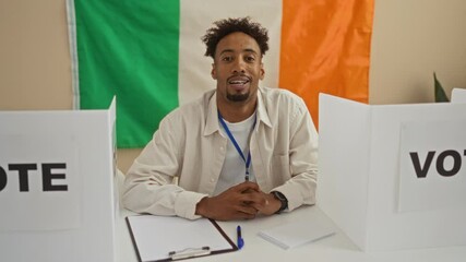 Handsome adult african american man with a beard sits in an indoor voting college room with irish flag in the background, surrounded by electoral materials.