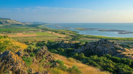 Breathtaking coastal landscape featuring a vast expanse of green fields, rocky hills, and a serene water body under a clear blue sky.