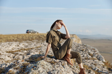 Woman sitting on rock with hands on head and car in background in a travel adventure moment