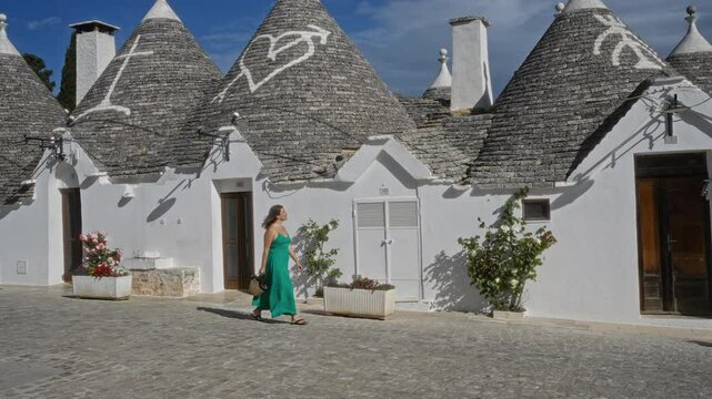 A young hispanic woman walks in front of traditional trulli houses in the old town of alberobello, puglia, italy, on a sunny day.