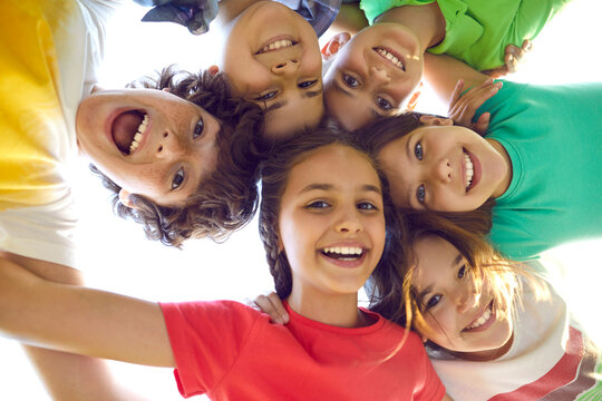 Happy childhood. Faces of funny joyful preteen friends who stand in circle and look down at camera. Bottom view of cute boys and girls in colorful summer casual clothes hugging and laughing outdoors.