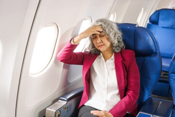 A mature woman sits by the window on a passenger plane, traveling abroad. She looks unwell, experiencing a headache, nausea, and vomiting, which dampens her travel excitement.