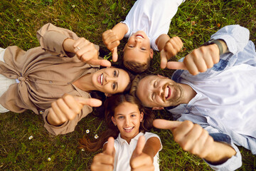 Top view of happy young family of four lying on green grass and showing thumb up sign in summer park looking at camera. Smiling parents having fun with kids in nature enjoying time together.