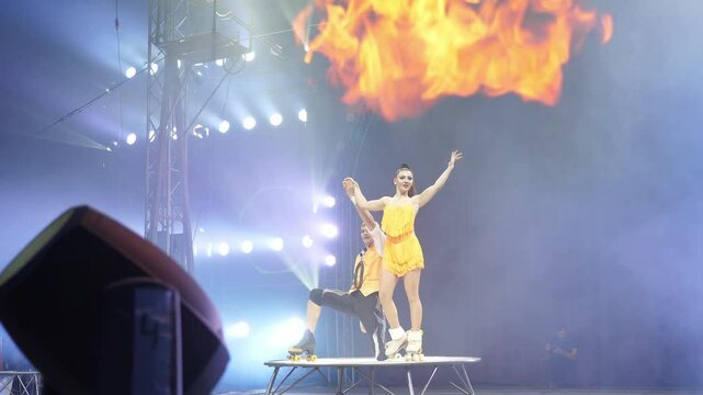 A pair of acrobats on roller skates in beautiful stage costumes perform in the circus arena, bowing at the end of the performance on a small stage. Concert illumination with smoke and fire.