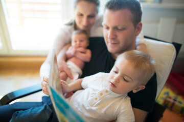 Obraz premium Nuclear family with neborn baby daughter reading book with older son. Mom and dad spending time with kids in room.