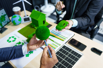 A meeting at an office desk focuses on teamwork with a house model and discussions on electric cars, solar cells, and green initiatives, emphasizing sustainability and environmental goals.