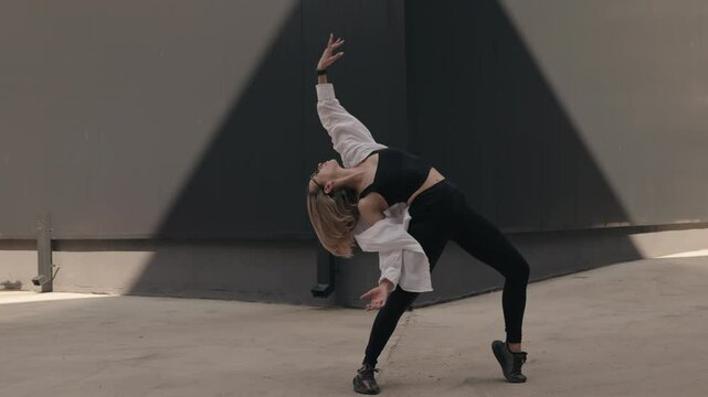 A young woman in a white shirt and black pants dances in an empty concrete lot. She is dancing in the sunlight near a large gray wall.