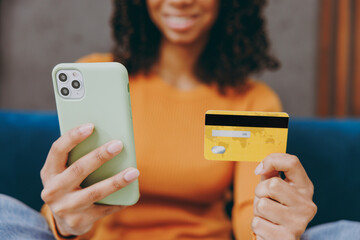 Little kid teen girl wear casual clothes use mobile cell phone credit bank card shopping sits on blue sofa couch stay home hotel flat rest relax spend free spare time in living room. Focus on device