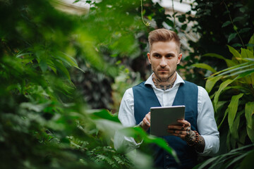 Handsome businessman with tablet, standing in botanical garden, in the middle of lush green...