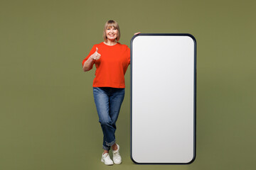 Full body smiling happy pretty woman 50s years old she wear orange t-shirt casual clothes big huge blank screen mobile cell phone smartphone with area show thumb up isolated on plain green background.