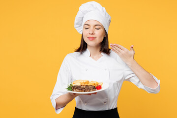 Young chef baker woman in white shirt black apron uniform toque chefs hat hold plate ready order dish steak, raise hand to face, feel food smell isolated on plain yellow background Cook food concept.