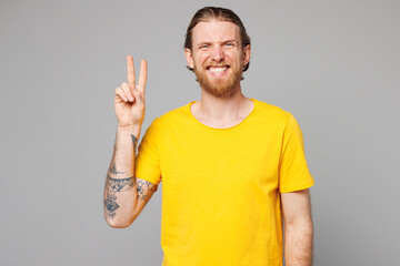 Young smiling happy fun joyful positive Caucasian man he wears yellow t-shirt casual clothes showing victory sign looking camera isolated on plain grey background studio portrait. Lifestyle concept.