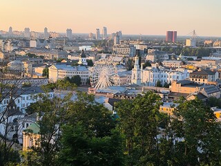 Top view of the city of Kyiv. Below is its old historical part. Cathedrals and old buildings. White Ferris wheel. In the distance you can see the river, high-rise buildings and bridges. Trees. Horizon