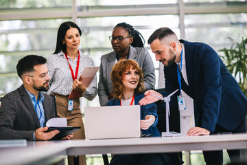 Multiracial business people talking about sustainable energy. They have windmill on a table and they are doing something on a laptop.