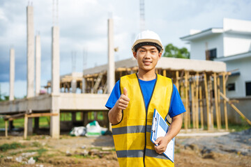 An architect, wearing a hardhat and safety vest, checks a laptop and house plan paper on a...
