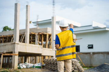 An architect, wearing a hardhat and safety vest, checks a laptop and house plan paper on a...