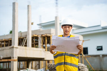 An architect, wearing a hardhat and safety vest, checks a laptop and house plan paper on a clipboard. Blueprints and construction documents lie nearby, emphasizing safety and precision in building.