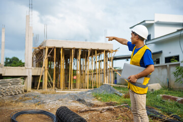 An architect, wearing a hardhat and safety vest, checks a laptop and house plan paper on a...