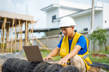 An architect, wearing a hardhat and safety vest, checks a laptop and house plan paper on a...