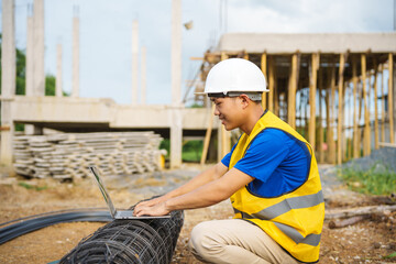 An architect, wearing a hardhat and safety vest, checks a laptop and house plan paper on a clipboard. Blueprints and construction documents lie nearby, emphasizing safety and precision in building.