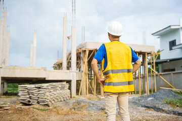 Fototapeta premium An architect, wearing a hardhat and safety vest, checks a laptop and house plan paper on a clipboard. Blueprints and construction documents lie nearby, emphasizing safety and precision in building.