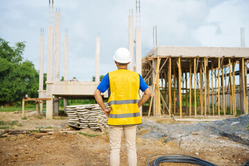 An architect, wearing a hardhat and safety vest, checks a laptop and house plan paper on a...