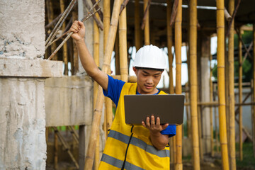 An architect, wearing a hardhat and safety vest, checks a laptop and house plan paper on a...