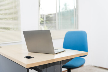 An empty desk in a minimalist office setting, featuring a blank workspace, large windows, a laptop, and a blue office chair against a white wall background, ideal for product montages. no people