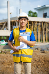 An architect, wearing a hardhat and safety vest, checks a laptop and house plan paper on a clipboard. Blueprints and construction documents lie nearby, emphasizing safety and precision in building.