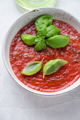 Bowl of tomato soup served with fresh green basil, vertical shot on a grey granite surface, elevated view, middle closeup