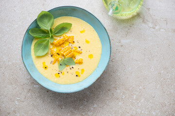 Sweet corn gazpacho served with fresh basil in a turquoise bowl, top view on a beige stone background, horizontal shot, copy space