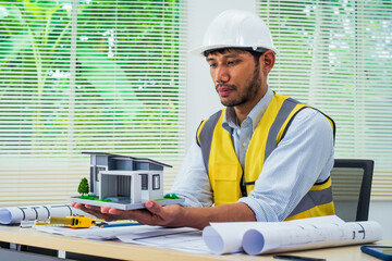 Asian architect works at a desk with house blueprints, checking plans on a laptop and clipboard,...