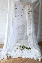 Bed with flowing curtains and garland flowers levitating, hanging flowers. Bedroom interior design.White Mattress for sleeping on the wooden floor with white linens in modern minimalist bright bedroom