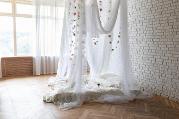 Bed with flowing curtains and garland flowers levitating, hanging flowers. Bedroom interior design.White Mattress for sleeping on the wooden floor with white linens in modern minimalist bright bedroom © stock_studio