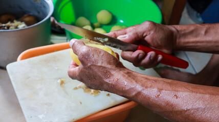 A close-up photo of a person peeling potatoes with a knife in a kitchen. The potatoes are yellow with a white cutting board in the kitchen.