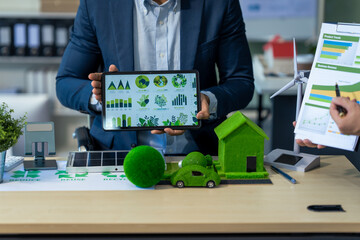 Two Asian businessmen in formal suits discuss ideas and suggestions at an office desk, focusing on green housing estates, city planning, renewable energy, and achieving net zero emissions by 2050.