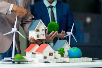 Two Asian businessmen in formal suits discuss ideas and suggestions at an office desk, focusing on green housing estates, city planning, renewable energy, and achieving net zero emissions by 2050.