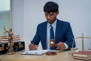 An Asian businessman in a formal suit works alone at a lawyer's desk, by golden scales, law books, leather bags, and a Lady Justice statue, emphasizing personal freedom and legal empowerment.