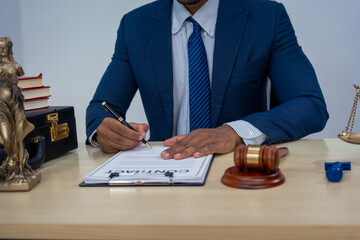 An Asian businessman in a formal suit works alone at a lawyer's desk, by golden scales, law books,...