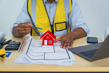 Asian architect works at a desk with house blueprints, checking plans on a laptop and clipboard,...