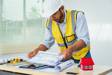 Asian architect works at a desk with house blueprints, checking plans on a laptop and clipboard,...