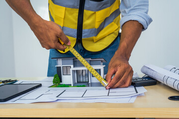 Asian architect works at a desk with house blueprints, checking plans on a laptop and clipboard,...