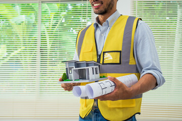 Asian architect works at a desk with house blueprints, checking plans on a laptop and clipboard,...