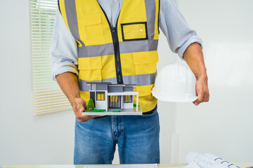 Asian architect works at a desk with house blueprints, checking plans on a laptop and clipboard, wearing a hardhat and safety vest, emphasizing precision and safety in office settings.