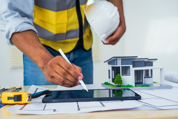 Asian architect works at a desk with house blueprints, checking plans on a laptop and clipboard, wearing a hardhat and safety vest, emphasizing precision and safety in office settings.