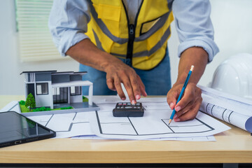 Asian architect works at a desk with house blueprints, checking plans on a laptop and clipboard,...