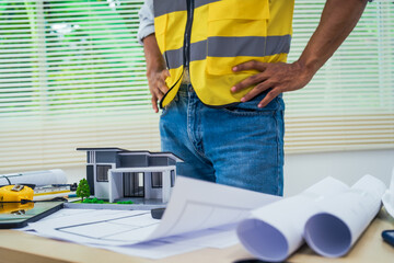 Asian architect works at a desk with house blueprints, checking plans on a laptop and clipboard,...