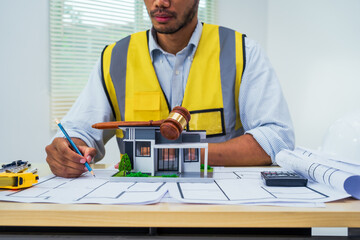 Asian architect works at a desk with house blueprints, checking plans on a laptop and clipboard,...