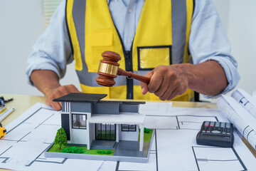 Asian architect works at a desk with house blueprints, checking plans on a laptop and clipboard,...